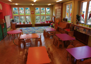 Na fotografii widok sali grupy czerwonej z oknami na wprost z leżącym dywanem i stołami w kolorach fioletowych i pomarańczowych dla dzieci